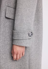 Zbliżenie ręki obok rękawa szarego wełnianego płaszcza z kieszenią klapową i detalem w postaci zapięciowej klamry.