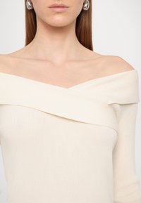Femme portant un pull crème côtelé décolleté et des boucles d'oreilles argentées en cercle, avec des cheveux bruns raides sur fond blanc.