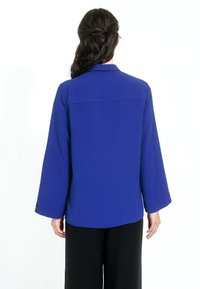 Blå blus med långa, vida ärmar, mjuk textur och krage. Har en ryggsömn och slutar under midjan. Kombineras med svarta byxor.