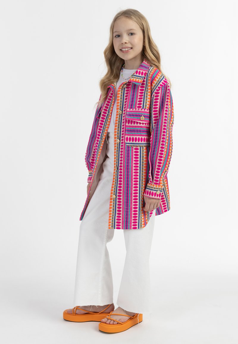 Spalvinga raštuota striukė su rožinėmis, oranžinėmis ir mėlynomis juostomis, derinama su baltomis platėjančiomis kelnėmis ir ryškiai oranžiniais platforminiais sandalais.