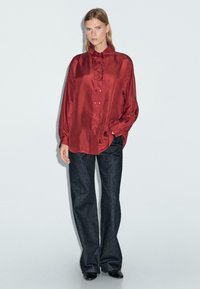 Rotes Satin-Hemd mit langen Ärmeln, hohem Kragen und Knopfleiste, kombiniert mit dunkelblauen Jeans im weit geschnittenen Stil und schwarzen Schuhen.