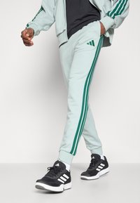 Ανοιχτό πράσινα φόρμα με πράσινες ρίγες στο πλάι, στενή σχεδίαση και ελαστικούς καρπούς. Φοριούνται με μαύρα αθλητικά παπούτσια με λευκές λεπτομέρειες.