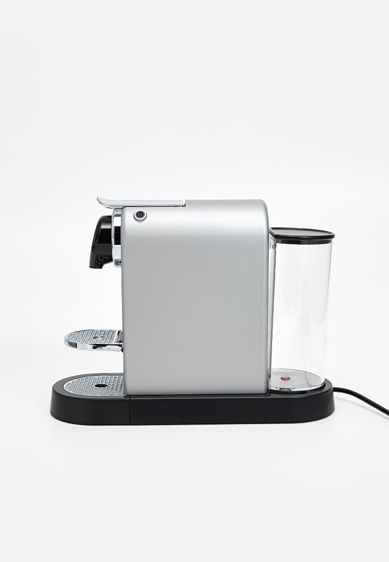 Machine à café argentée au design épuré, réservoir d'eau en verre, base noire, accents en chrome et plateaux d'égouttage texturés. Bouton d'alimentation visible.