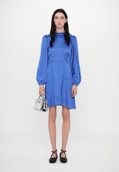 Mėlyna satino suknelė su tekstūruotu raštu, ilgomis pūstomis rankovėmis, rauktu iškirpte ir platėjančia apačia. Modelis laiko mažą sidabrinę rankinę.