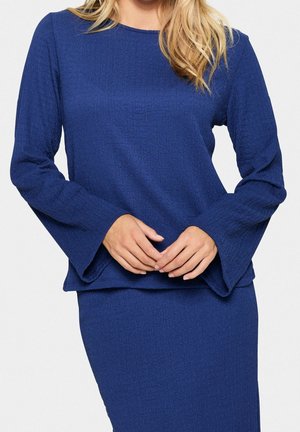 Woman wearing a textured royal blue long-sleeve top with flared sleeves and matching fitted skirt, hands lightly clasped in front.