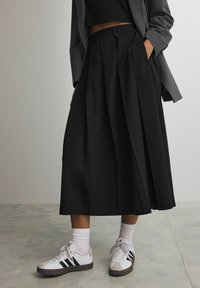 Czarna plisowana spódnica midi z wysokim stanem i bocznymi kieszeniami, w połączeniu z białymi tenisówkami z czarnymi paskami i prążkowanymi białymi skarpetkami.