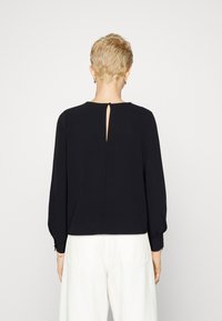 Black long-sleeve top with a round neckline, featuring a decorative keyhole opening at the back and pleated sleeves. Smooth fabric texture.