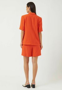 Oranssi lyhythihainen paita ja shortsit, ribbitekstuuri, rento istuvuus, kauluskantti ja takasauman yksityiskohta. Jalkineina on lohkarekorot sandaalit.