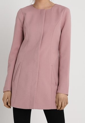 Mujer con abrigo ajustado de manga larga en color rosa polvoriento, con cierre frontal oculto y bolsillos laterales en ángulo, combinado con pantalones negros.