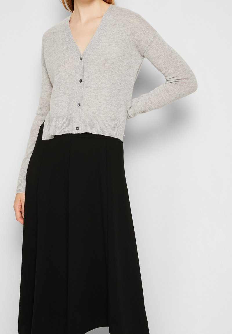 Helle graue, verkürzte Strickjacke mit tiefem V-Ausschnitt, Knopfleiste und gerippten Bündchen, kombiniert mit einem langen schwarzen Rock. Glatte Textur und schlichtes Design.
