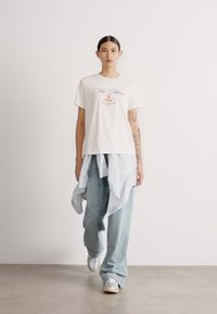 White t-shirt with printed design, paired with light blue wide-leg jeans and a striped, tied pullover. Silver sneakers complete the look.