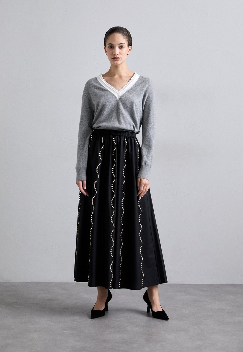 Grauer Strickpullover mit V-Ausschnitt und weißem Spitzenbesatz, kombiniert mit einem schwarzen Maxirock mit wellenförmigen weißen Pailletten und einem elastischen Bund.