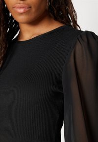 Schwarzes Top mit gerippter Textur, Rundhals-Ausschnitt und transparenten Puffärmeln. Der Stoff ist an den Schultern glatt und fließend.