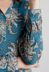 Blå skjorte med brunt og hvidt paisle mønster, knaplukninger ved håndleddet, med den ene arm bøjet og hånden holdende om den modsatte albue.