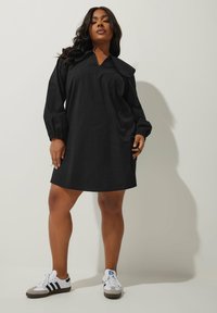 Svart oversized klänning med v-ringning och volanger, långa ärmar och mjuk textur, bärs med vita och gråa träningsskor.