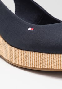 Primo piano di una scarpa piatta in tessuto blu navy con suola platform intrecciata beige e piccolo logo ricamato rosso, bianco e blu.