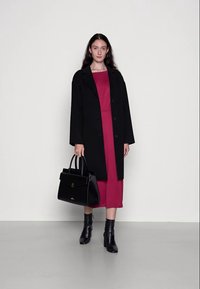 Casaco de lã preto sobre um vestido bordô, combinado com uma bolsa de couro preta e botins pretos, com texturas suaves e uma forma estruturada.
