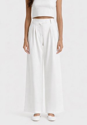 Donna che indossa pantaloni bianchi a vita alta e gamba larga, top corto senza maniche e scarpe bianche basse, in piedi davanti a uno sfondo semplice.