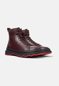 Stivali alla caviglia in pelle bordeaux con suole in gomma nera e rossa, dotati di un design alto, punta rotonda e lacci frontali per una vestibilità regolabile.