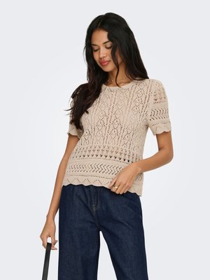 Femme aux longs cheveux foncés portant un haut beige à manches courtes en crochet et un jean large bleu foncé, tenant une sangle noire dans la main gauche.