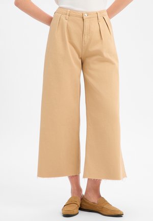 Beige weit geschnittene, gekürzte Hose mit rohem Saum, getragen mit braunen Slip-on-Loafern bei einer Person, die vor weißem Hintergrund steht.