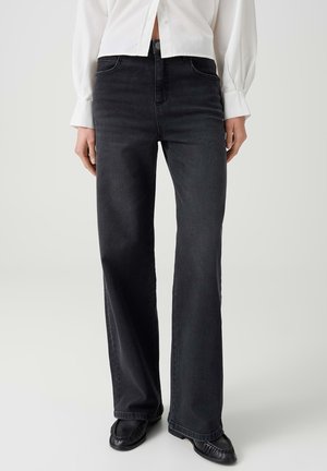 Person trägt schwarze Jeans mit hoher Taille und geradem Bein, kombiniert mit schwarzen Schuhen und einem weißen langärmligen Hemd mit Knöpfen.