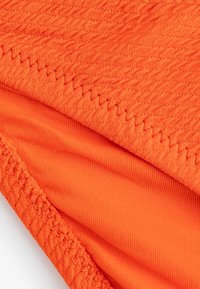 Tessuto arancione con sezioni texture e lisce. Presenta cuciture visibili lungo i bordi, enfatizzando il colore vivace del tessuto.