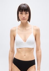 Γυναίκα με κοντά σκούρα μαλλιά που φοράει ένα λευκό μπραλέτο και μαύρο εσώρουχο, στέκεται μπροστά σε ένα απλό ανοιχτό γκρι φόντο.