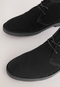 Schwarze Wildleder-Schnürschuhe mit Ziernähten und Gummisohlen auf hellem Hintergrund.