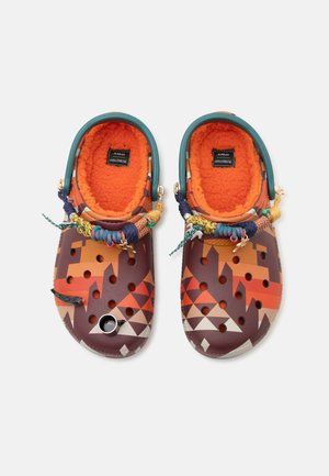 Paire de sabots à enfiler à motifs avec doublure polaire orange, brides vertes au talon et lacets tressés multicolores avec breloques sur fond blanc.