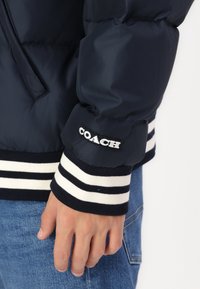Námornícka pufrová bunda s kontrastným bielym a námorníckym pruhovaným rebrovaným manžetom. Na rukáve sa nachádza logo "COACH". Hladký, lesklý povrch.