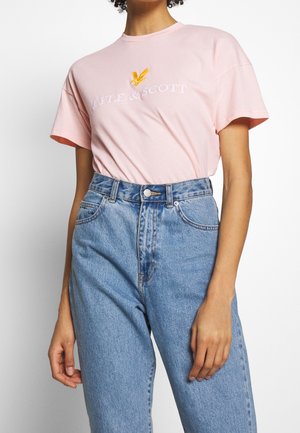 Persoon draagt een lichtroze Lyle & Scott T-shirt met een geel adelaar-logo, ingestopt in een hooggetailleerde blauwe spijkerbroek.