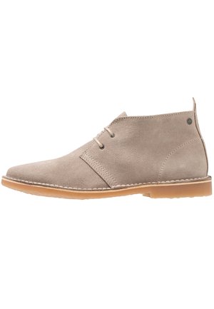 Beżowy zamszowy, sięgający do kostki but desert boot z dwoma oczkami, beżowymi sznurówkami, jasnobrązową gumową podeszwą i pętelką z tyłu przy pięcie.