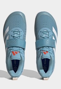 Chaussures de sport bleu clair avec un tissu texturé, des rayures blanches et un design de sangle rembourrée. Présente des accents rouges et un renfort au niveau du talon.