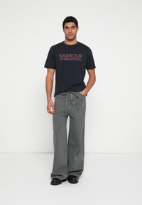 Tricou negru cu textul „BARBOUR INTERNATIONAL” imprimat cu litere îndrăznețe bordeau, asortat cu pantaloni gri cu picioare largi și pantofi negri.