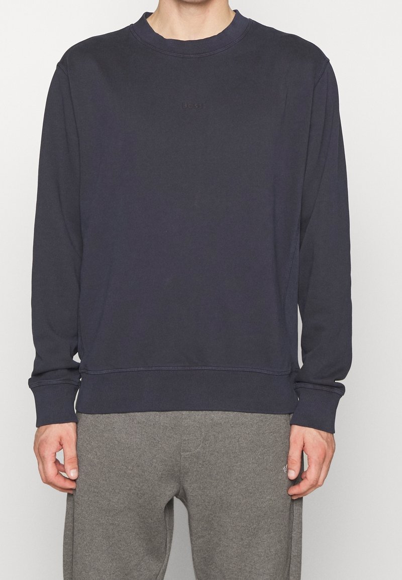 Mörkblå sweatshirt i ett mjukt tyg; har rundad halsringning, ribbade ärmslut och en liten tonal logotyp på bröstet. Matchas med gråa sweatpants.