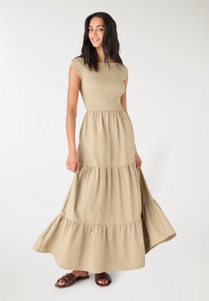 Donna con capelli lunghi e scuri che indossa un abito maxi a balze beige e sandali scivolati marroni, in piedi davanti a uno sfondo bianco semplice.