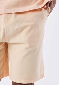 Pantaloni corti in cotone pesca chiaro con una texture liscia, dotati di tasche laterali e orli cuciti. La vita presenta un dettaglio elastico discreto.