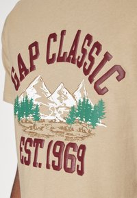 Beżowa bawełniana koszulka z grafiką góry i drzewa. Na maroonowym tle napis "GAP CLASSIC" oraz "EST. 1969." Miękka tekstura, swobodny design.