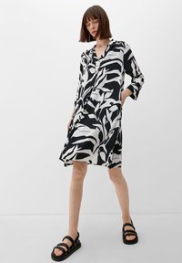 Schwarz-weiß gemustertes Kleid mit langen Ärmeln und Knopfleiste. Der Stoff ist glatt, und das Design zeigt große abstrakte Formen.