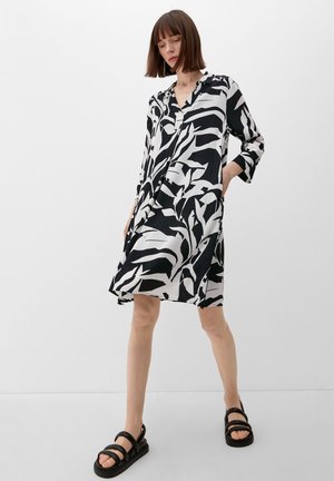 Robe à motifs noir et blanc avec des manches longues et une patte de boutonnage. Le tissu est lisse et le design comporte de grandes formes abstraites.