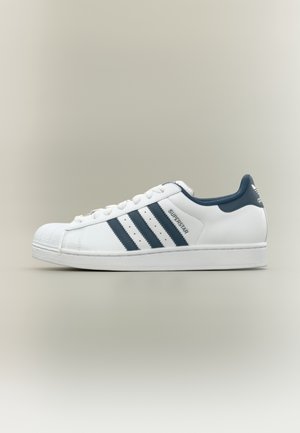Weißer Adidas Superstar Sneaker mit schwarzen Streifen, Shell-Toe und schwarzem Kragen, seitlich auf neutralem Hintergrund dargestellt.