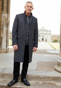 Hombre mayor con cabello corto y canoso, vestido con un abrigo gris oscuro, pantalones negros y zapatos negros, se encuentra frente a un edificio histórico.