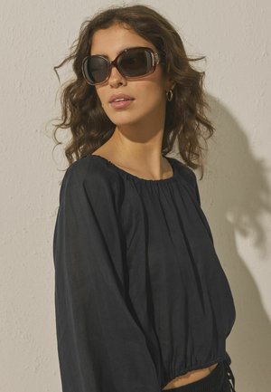 Femeie cu păr ondulat, purtând ochelari de soare mari și întunecați, cercei rotunzi aurii și o bluză neagră lejeră, în fața unui perete texturat deschis la culoare.