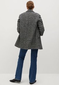 Osoba stojící zády, oblečená v šedém sako se vzorem kostky rybí kost, modrých džínách a černých balerínách, s vlasy staženými do drdolu, na jednobarevném pozadí.