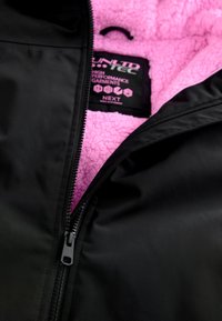 Veste noire partiellement dézippée pour montrer la doublure polaire rose et l’étiquette de vêtement avec la marque et les détails d’entretien à l’intérieur du col.