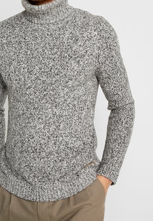 Mężczyzna w szaro-czarnym melanżowym golfie z rękami w kieszeniach beżowych spodni.