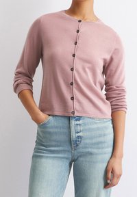 Cardigan léger rose à manches longues, col rond et boutons foncés sur le devant. Porté sur un jean en denim bleu clair.
