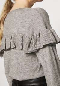 Femme portant un pull en tricot gris avec des volants sur le haut du dos et un pantalon en cuir noir.