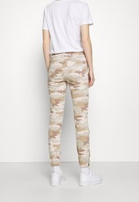 Joggeurs camouflage en tons beige et marron clair, avec une taille élastique, des jambes fuselées et deux poches arrière. Portés avec des baskets blanches.